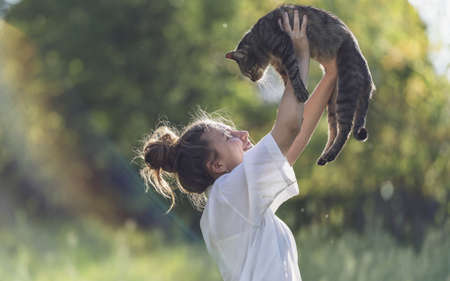 in the photo is a girl holding a tabby cat over her head. Cat with girl looking into each other's eyes. It can be seen that they have a friendly relationship. The girl laughs at the cat.の写真素材