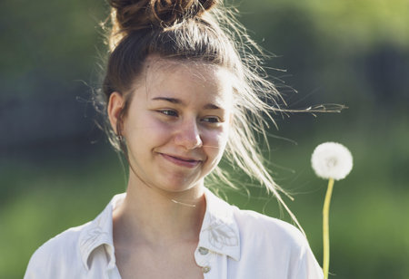 The girl holds a dandelion in her hand and looks at it with a smile. She is about to blow, but first examines the flower. It is a beautiful sunny day and the girl is outside in nature.の写真素材