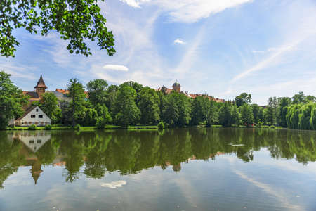 in the photo there is a pond surrounded by trees and buildings are displayed. The reflection of the world around is reflected from the surface. The sky is blue and it's beautifulの写真素材