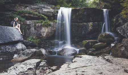The photo shows a blonde woman turned and looking towards the waterfall. A woman sits on a rock on the left side and waterfalls flow in the middle and on the right side of the photoの写真素材