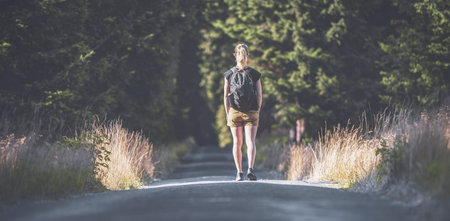 The photo shows a blonde woman facing the direction of the forest, where the path she is walking leads to. The woman has a backpack on her back and is dressed in short travel shortsの写真素材