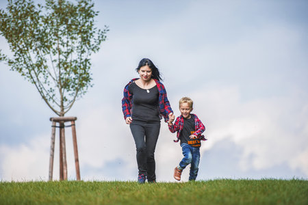 A little boy in a checkered shirt and blue pants with his mom are walking on top of a meadow near the horizon. Behind them he saw the sky dotted with clouds. small trees grow nextの写真素材