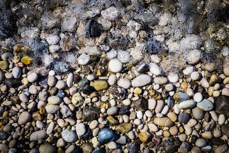 Background of multicolored sea pebbles on the beach in sea foam. Small color wet stones gravel texture background.の写真素材