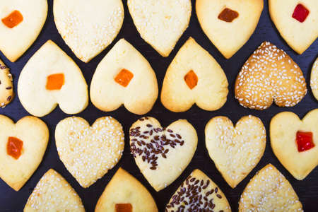Background of homemade heart shaped cookies with sesame seeds, flax seeds, candied fruit baked for Valentine day 14 February on dark brown table. Top view. Love sweet backdrop.の写真素材