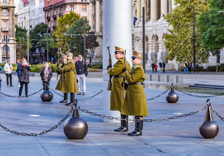 Budapest, Hungary - 10 of October 2019: Soldiers guard of honor in traditional green greatcoats near National Hungarian Flag on top of tall steel flagpole on the Kossuth Lajos Square near Parliament building.のeditorial素材