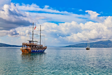 Vintage mast wooden sailing ship for sea tours in Saranda gulf, Albania with red state albanian flag with black double-headed eagle. Small yacht and Corfu island on the horizonのeditorial素材