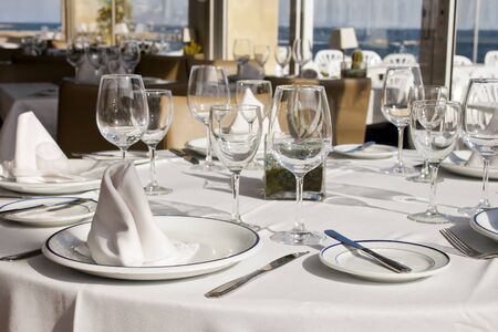 Table crockery setting in a restaurant next to the sea. Glasses, plates, cutlery and napkins on a tablecloth.の写真素材