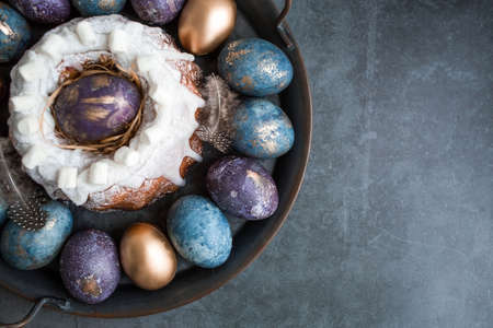 Easter card with a copy of the place for the text. Purple, blue and golden eggs with a cake on a dark background. The purple hue trend of 2022 is very peri. Natural dye karkade tea. top view.の写真素材