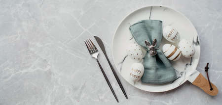 Banner. table settings. A fashionable marble plate with a rabbit on a napkin, Easter eggs and feathers on a gray background. top view. Happy Easter holiday concept for cafes and restaurants.の写真素材