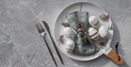 Banner. table settings. A fashionable marble plate with a rabbit on a napkin, Easter eggs and feathers on a gray background. top view. Happy Easter holiday concept for cafes and restaurants.の写真素材