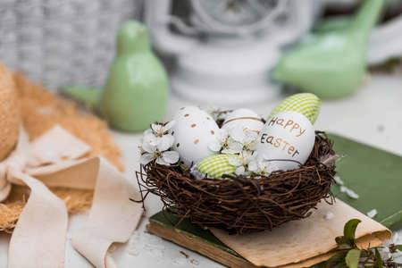 Banner. White-green eggs, cherry blossoms in a nest, an old book and porcelain birds on a white wooden table. A card festive for happy Easter Day 2022 in vintage style.の写真素材