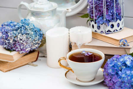 A visual content. Still life in vintage style. A mug with a drink, an old book, a cage, candles and hydrangea flowers in the garden on a white wooden table. The concept of a tea ceremony.の写真素材