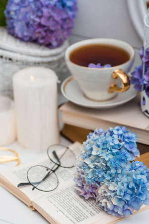 A visual content. Still life in vintage style. A mug with a drink, an old book, a cage, candles and hydrangea flowers in the garden on a white wooden table. The concept of a tea ceremony.の写真素材
