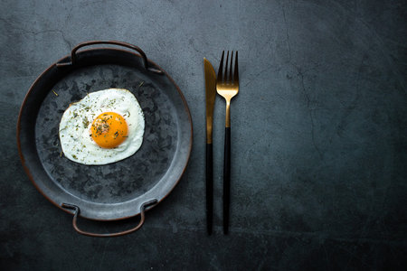 Homemade fried egg in a vintage pan and stylish cutlery on a dark background. view from above. Copy...