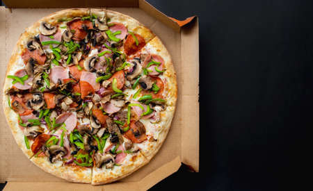 Traditional italian pizza with pepproni sausage, bacon, mushrooms, tomatoes, green pepper and cheese on a black background. view from above. Copy space for text. Restaurant menu concept.Takeaway food.の写真素材