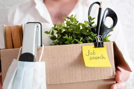 Dismissal from work and job search. Image of an office worker in a medical mask in formal attire. A man holds a box with his things, on a light background. The global crisisの写真素材