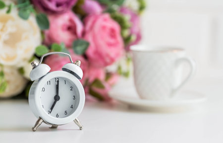 White retro alarm clock, flowers and a cup of coffee on the background of a white brick wall. The...