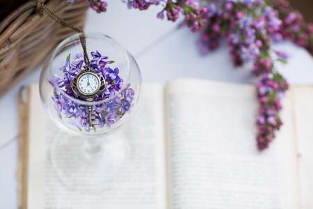 Beautiful still life, spring card.Lilac and vintage antique clocks lie in a glass goblet surrounded by flowers on a white background.The clock is 7 in the morning. Good morning concept. Moscow, May 2020の写真素材