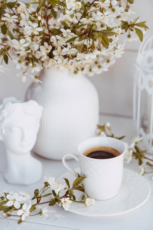 A beautiful postcard. A white coffee cup with a saucer, a statuette, candles and a vase with a bouquet of cherry blossoms. beautiful still life. springtime. The concept of "Good morning".の写真素材
