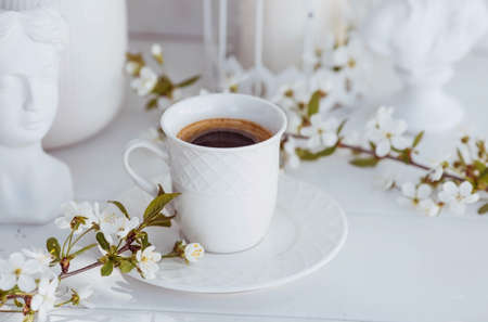 A beautiful postcard. A white coffee cup with a saucer, a statuette, candles and a vase with a bouquet of cherry blossoms. beautiful still life. springtime. The concept of "Good morning".の写真素材