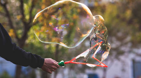 A man's hand makes big huge bright soap bubbles against the background of trees. Soap bubble show. Summer children's active recreation. Abstract blurred background.の写真素材