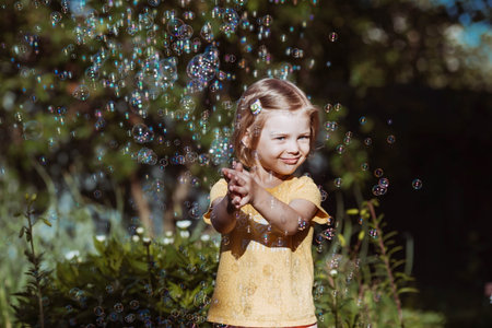 Cute happy girl playing with soap bubbles on a sunny summer evening. A child in the fresh air, in...