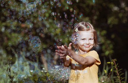 Cute happy girl playing with soap bubbles on a sunny summer evening. A child in the fresh air, in...
