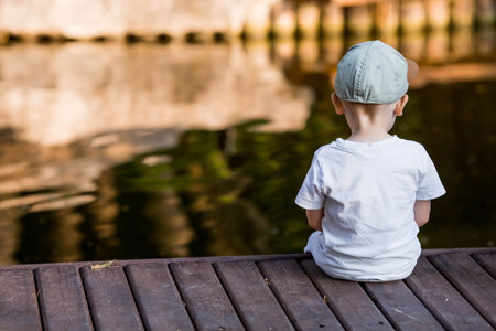 A little boy in a cap sits on a wooden pier and looks at the waterの写真素材