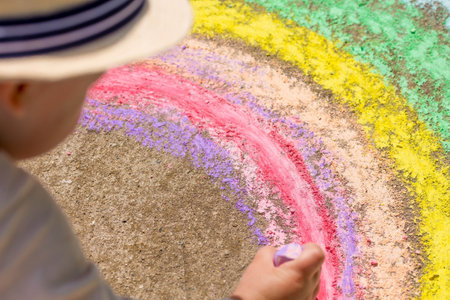 A happy child draws a rainbow with chalk on the asphalt outdoors. Portrait of a boy drawing with rainbow colored chalk on a sunny summer day. Creative development of children.の写真素材