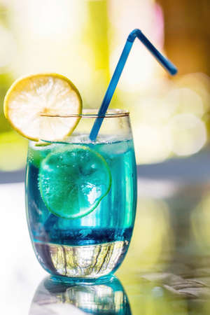 A glass of delicious blue cocktail blue lagoon and yellow lemons on the background of nature. Alcoholic cocktail juicy fruit blue with curacao liqueur, ice cubes and lemon slices.の写真素材