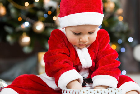 A beautiful little boy in a Santa Claus costume laughs merrily and opens a gift against the background of Christmas lights. The concept of Christmas celebration. new year holiday.の写真素材