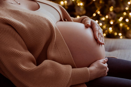 A young pregnant woman holding her tummy against the background of a Christmas tree. close-up. A woman is preparing to become a mother. A gift for the new year. Pregnancy and childbirth.の写真素材