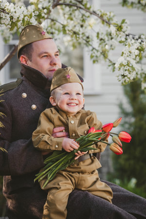 A boy in a military uniform with a bouquet sits on a soldier's lap against a background of cherry blossoms. The concept of the Great Victory Day of 1941-1945 and Defender of the Fatherland Day.の写真素材