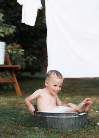 A little boy bathes, washes in an iron basin. A happy child splashes in the water. Vintage. The concept of child hygiene. Soap bubbles, a bubble bath.の写真素材