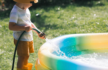 A little boy is playing with a garden hose in the backyard on a sunny day. A preschool child pours an inflatable pool. The concept of raising a child and teaching him to work.の写真素材