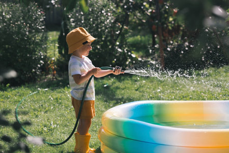 A little boy is playing with a garden hose in the backyard on a sunny day. A preschool child pours an inflatable pool. The concept of raising a child and teaching him to work.の写真素材