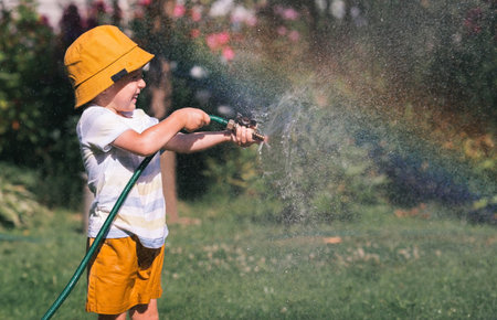 A little boy is playing with a garden hose in the backyard on a hot sunny day. A preschool child is watering flowers in the garden. The concept of raising a child and teaching him to work.の写真素材