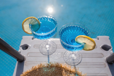 A glass of delicious blue cocktail Blue Lagoon and yellow lemons on the background of the pool. Alcoholic cocktail juicy fruit blue with curacao liqueur, ice cubes and a slice of lemon.の写真素材