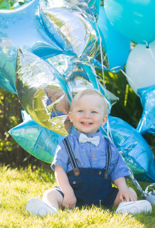 A beautiful little boy kid gentleman in a retro suit with suspenders and a bow tie is sitting on a green lawn. Children's party with balloons happy birthday, 1 year.の写真素材