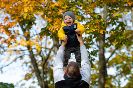 Cute little boy playing with dad outdoors. A happy child is tossed up in an autumn Park. The kid laughs merrily. Portrait of a boy. Autumn fashion. Stylish child on the street.の写真素材