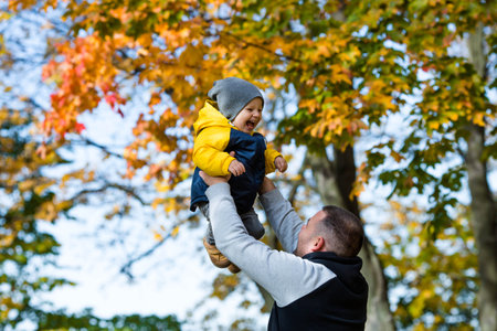 Cute little boy playing with dad outdoors. A happy child is tossed up in an autumn Park. The kid laughs merrily. Portrait of a boy. Autumn fashion. Stylish child on the street.の写真素材