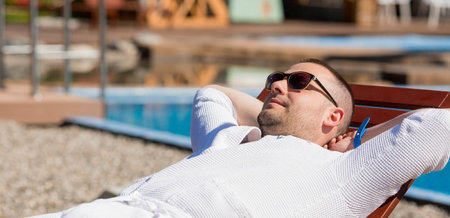 Portrait of a relaxed young man in sunglasses and a white coat, sunbathing by the pool with his...