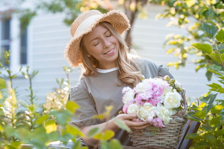 Beautiful blonde in a green summer garden. A girl in a straw hat with a bouquet of white peonies and pink carnations.の写真素材