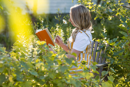 Beautiful blonde woman in a green summer garden on a chaise longue. A girl in a yellow sundress and with a bouquet of flowers reads a book. The concept of a country holiday in nature.の写真素材