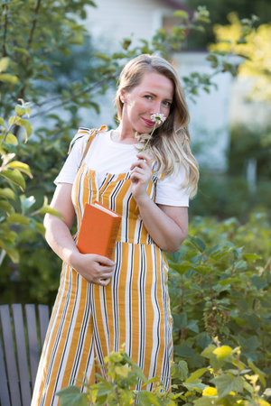 Beautiful blonde woman in a green summer garden on a chaise longue. A girl in a yellow sundress and with a bouquet of flowers reads a book. The concept of a country holiday in nature.の写真素材