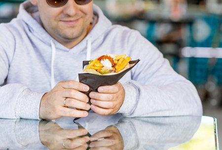 Fast street food and lifestyle. A young man eats harmful delicious gyros in a fast food restaurant in the fresh air. The man smiles and is happy.の写真素材