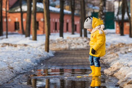 A small child in yellow rubber boots and a jacket runs through puddles, has fun, plays and launches paper boats. spring break photo. It's springtime.の写真素材