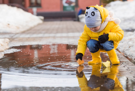 A small child in yellow rubber boots and a jacket runs through puddles, has fun, plays and launches paper boats. spring break photo. It's springtime.の写真素材