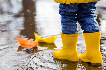 It's springtime. A small child in yellow rubber boots jumps through puddles, plays and launches paper boats on the water.. Photo of spring and autumn holidays. The concept of spring.の写真素材
