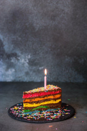 Rainbow birthday cake decorated with a festive candle and confetti. confectionery. Multicolored dessert in the color of the rainbow on a black background.の写真素材
